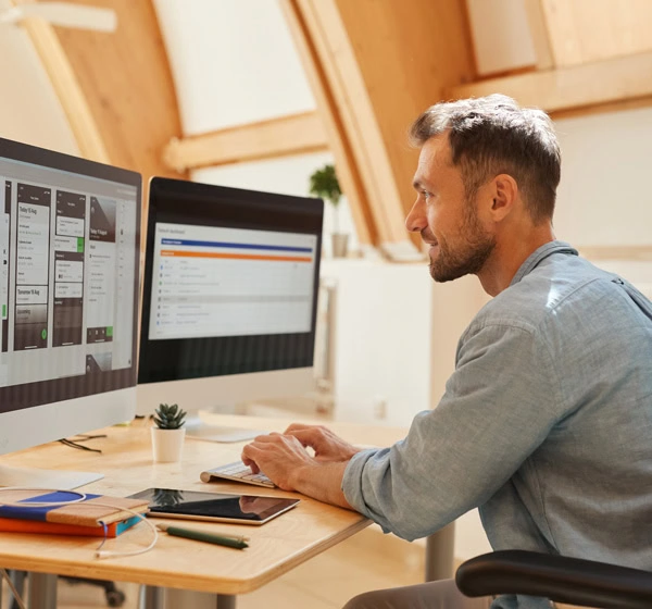 A man in a light blue shirt works on a computer at a desk with two monitors displaying software and data related to sourcing strategy in a bright office space.