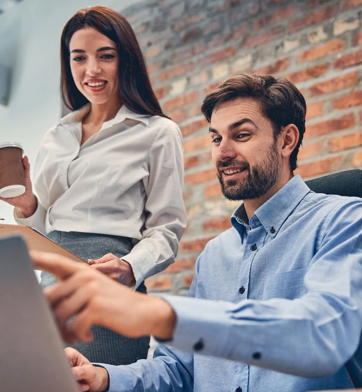 A man sitting at a desk points at a laptop screen, guiding the woman beside him who holds a coffee cup and clipboard. Both appear focused on choosing the right ERP solution, set against a backdrop of an exposed brick wall, choosing the right ERP solution
