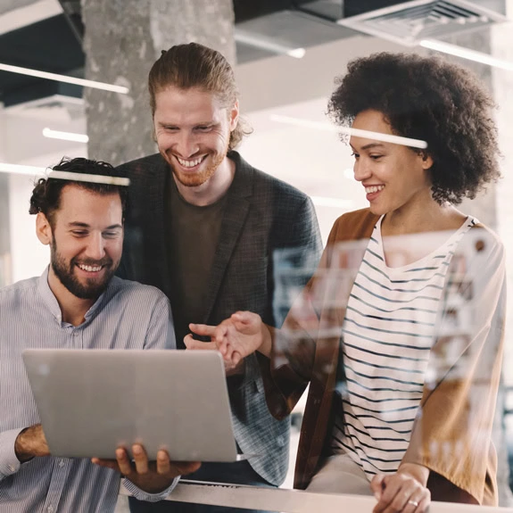 Three people in an office setting look at a laptop screen together, smiling and discussing their sourcing strategy. The atmosphere is collaborative and positive, highlighting their teamwork.