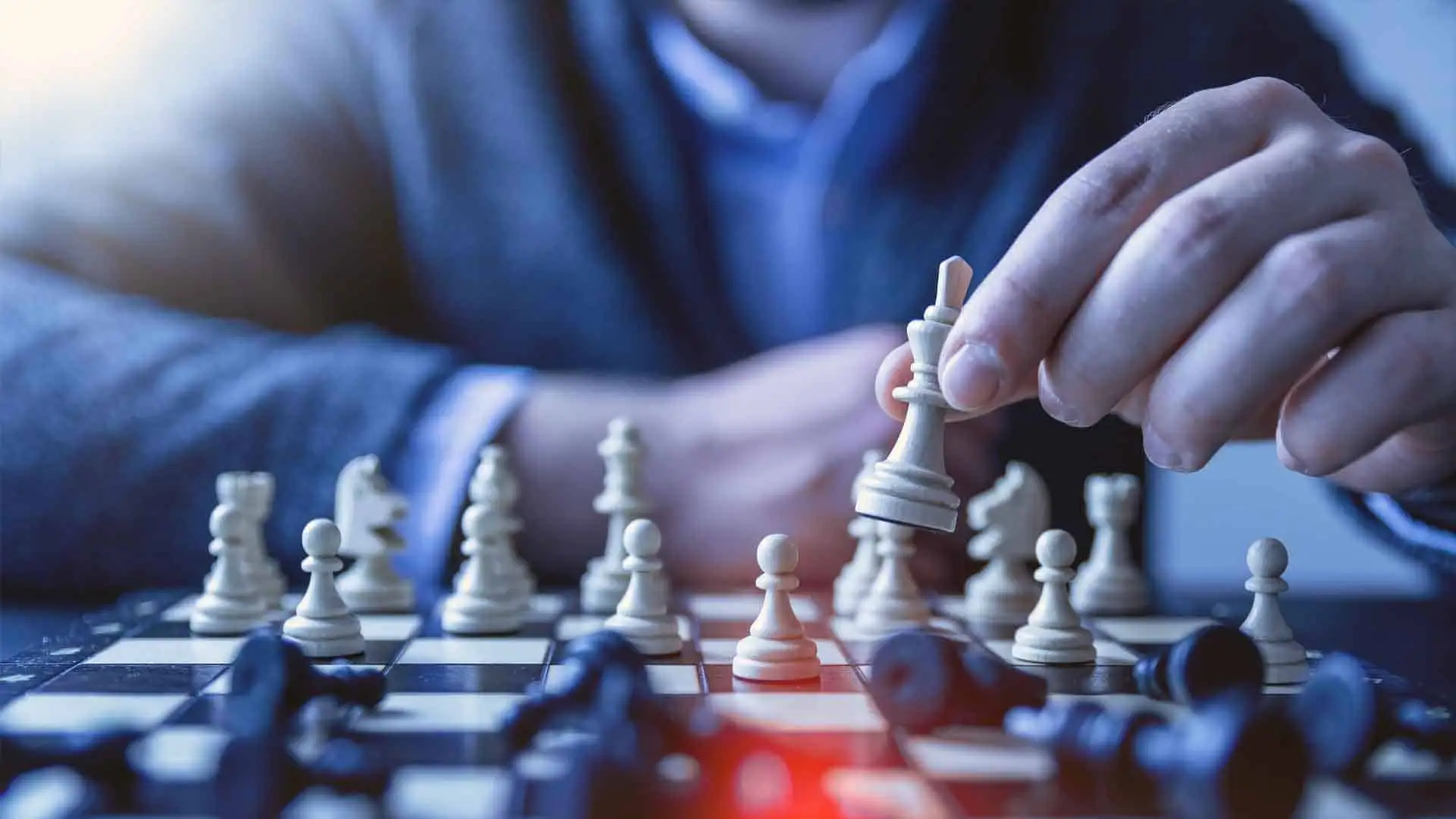 A person strategically holds a chess piece above a chessboard, surrounded by fallen pieces, much like refining a sourcing strategy in the midst of competition.
