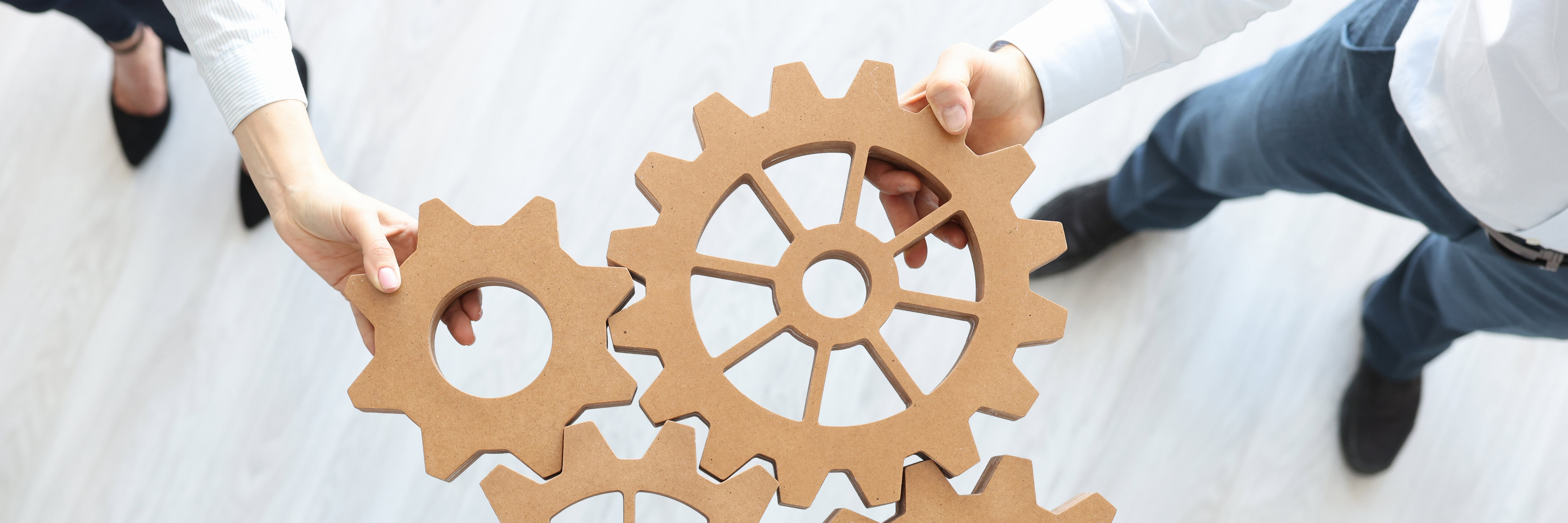 Two people hold and connect four large cardboard gears together over a light-colored floor, symbolizing teamwork or collaboration.